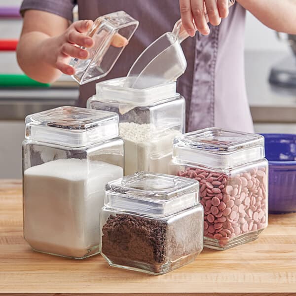 A woman pouring flour from a measuring cup into a clear glass jar.