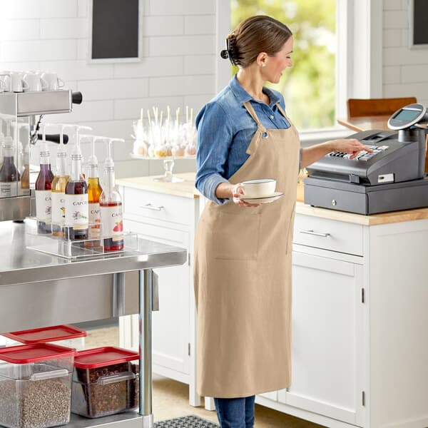 A woman in an Acopa Ashville natural linen bib apron holding a cup and a coffee cup.