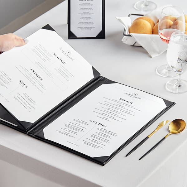 A person using a spoon and knife to open an Acopa Prime black vinyl menu on a table.
