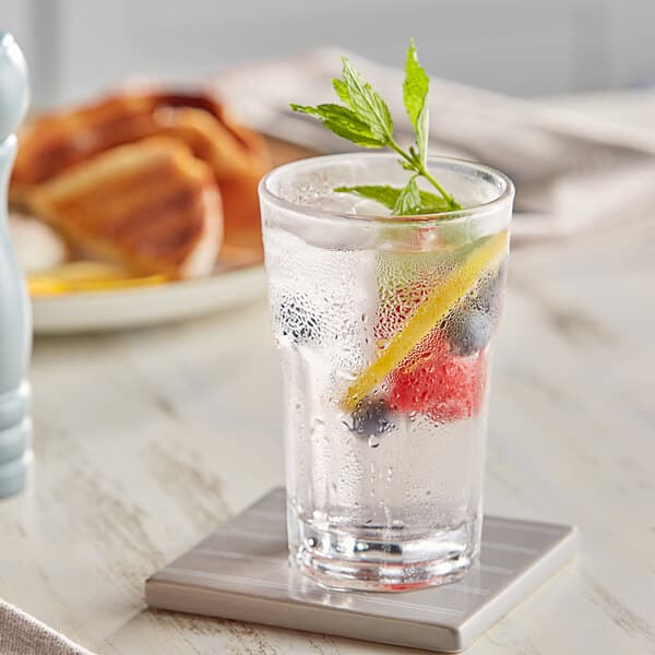 A clear highball glass filled with ice water, garnished with fruit slices and mint, placed on a coaster.
