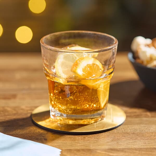 An Acopa Memphis old fashioned glass of whiskey with ice and a lemon slice on a table.