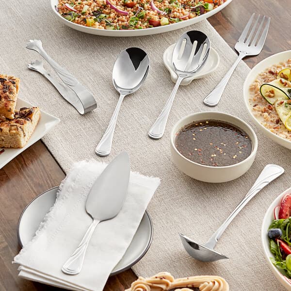 Acopa stainless steel serving utensils on a table with plates of food and silverware.