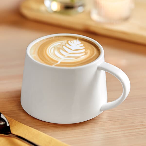 An Acopa bright white porcelain coffee cup filled with coffee with a leaf design in the foam.