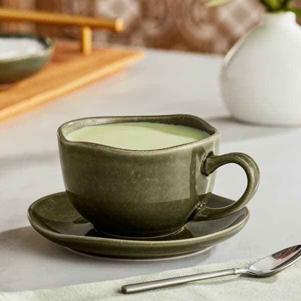 A green Acopa Highlands china cup with liquid in it on a table with a saucer and a spoon.
