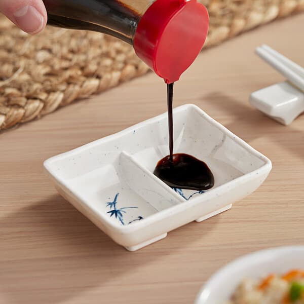 A hand pouring soy sauce into an Acopa Biru rectangular divided sauce dish.