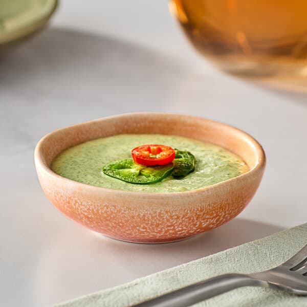A bowl of green sauce with a red pepper on top in an Acopa Primrose Peach sauce cup.