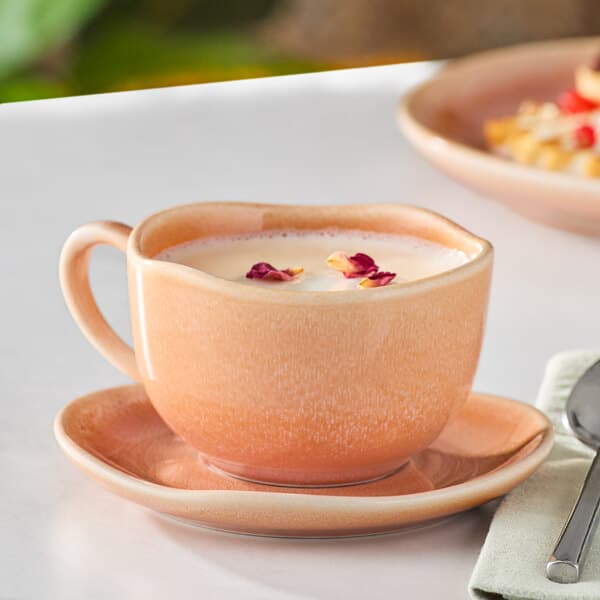 An Acopa Primrose Peach China cup of milk with rose petals on a saucer.