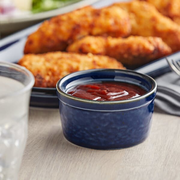 A blue and white Acopa Keystone porcelain ramekin filled with red sauce on a table with a plate of chicken nuggets.