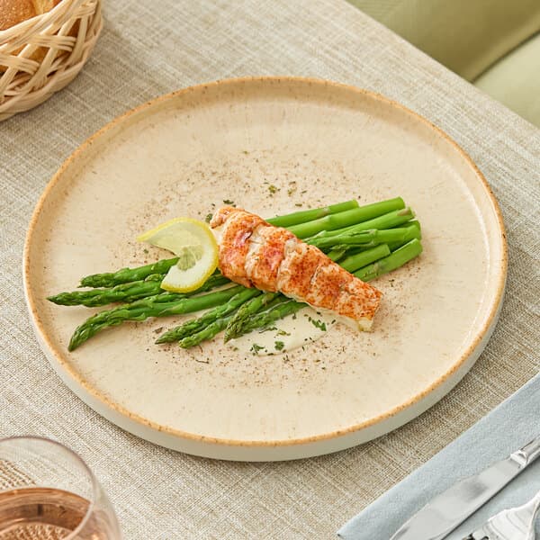 A round beige porcelain plate with a serving of asparagus, lobster, and a lemon wedge.