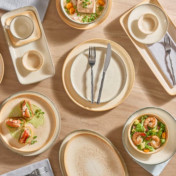 A set of beige and gray porcelain dinnerware, including plates, bowls, cups, and butter dishes, arranged on a wooden table.