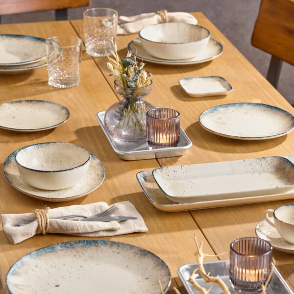 A small square gray porcelain butter dish placed on a wooden dining table set with various dinnerware and glassware.