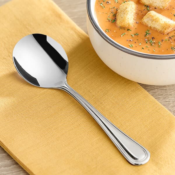 An Acopa Edgeworth stainless steel bouillon spoon on a napkin next to a bowl of soup.