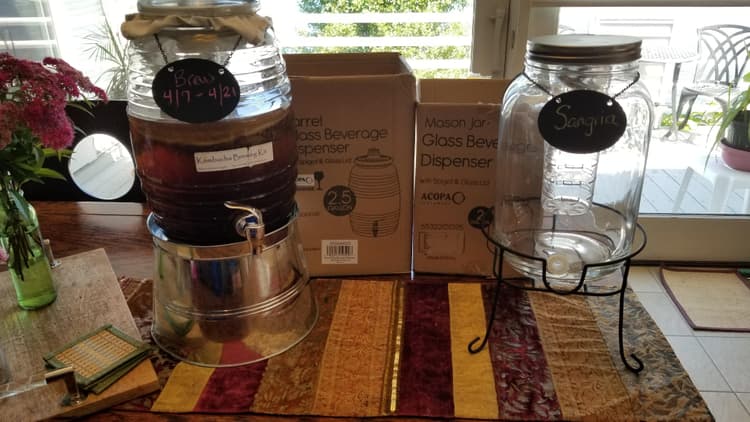Acopa 2 Gallon Mason Jar Glass Beverage Dispenser with Infusion Chamber, Chalkboard Sign, and Black Stand pictured on left is next to the 2.5g with other style stand I like both and it's great they (stands infusers and chalk tags) are interchangeable