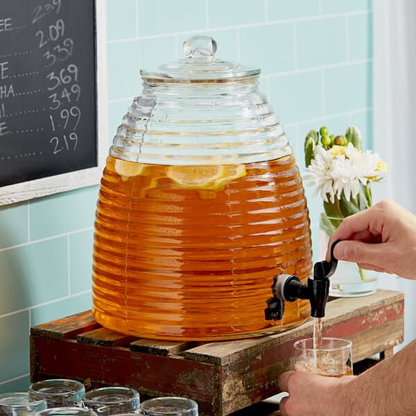 A hand pouring a drink from an Acopa glass beverage dispenser into a glass with a lemon slice.