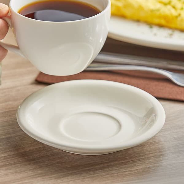 An Acopa ivory stoneware saucer under a cup of coffee on a table.