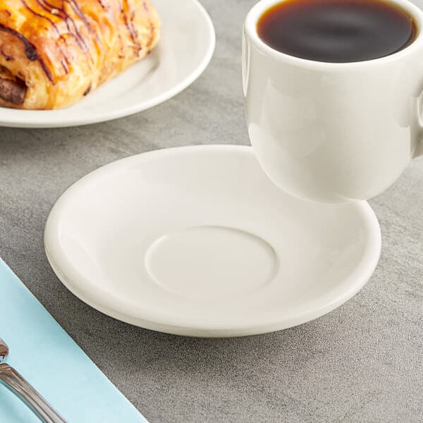 An Acopa ivory saucer with a cup of coffee on a table next to a plate of food.