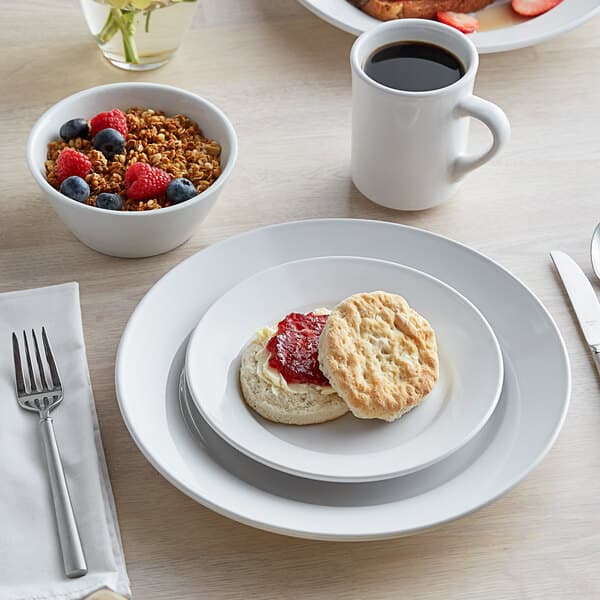 A white Acopa stoneware plate with a bowl of cereal, berries, and a biscuit with jam on it.
