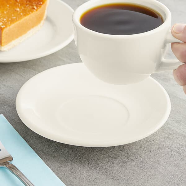 An Acopa ivory saucer holding a cup of coffee on a table with a fork and knife.