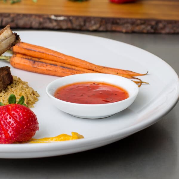 A white plate of food with a sauce and vegetables with a white round sauce cup on it.