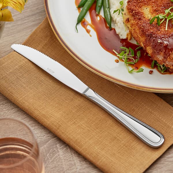 An Acopa Edgeworth stainless steel dinner knife on a plate with meat, green beans, and sauce.