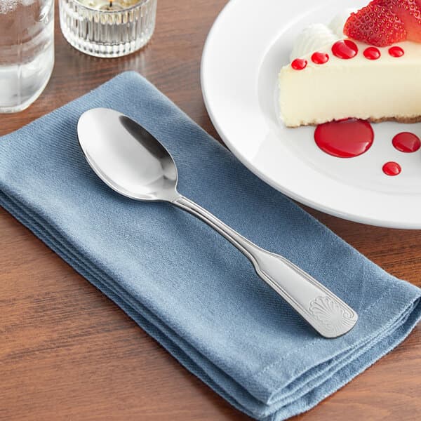An Acopa stainless steel spoon on a blue napkin next to a plate of cheesecake.