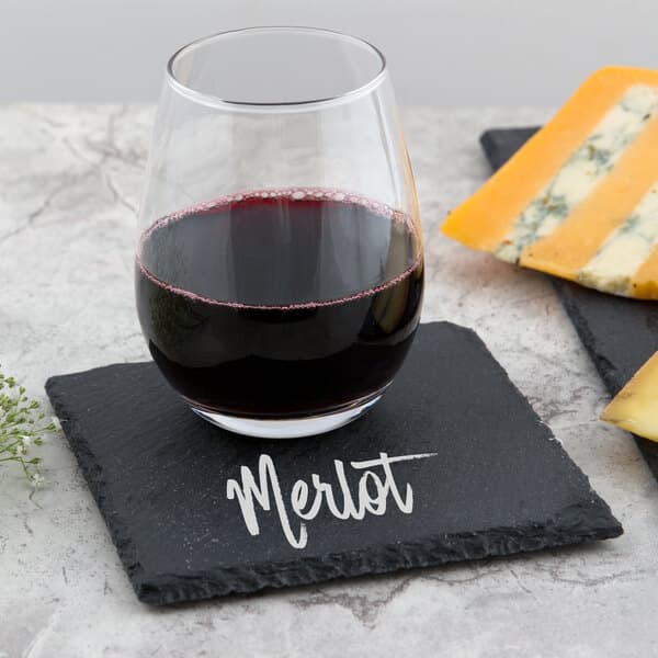 A glass of wine on a slate coaster next to a cheese plate with the word "merlot" written in chalk.