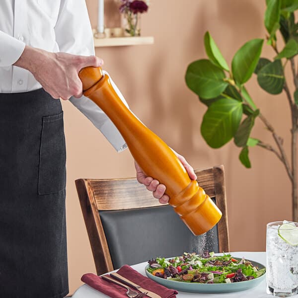 A person holding an Acopa matte brown wooden pepper mill over a plate of salad.