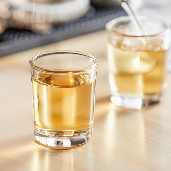 An Acopa shot glass filled with yellow liquid on a table with another filled shot glass.