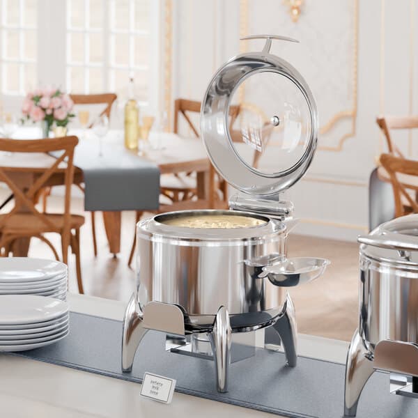 An Acopa stainless steel soup chafer with a glass lid on a table with plates and chairs.