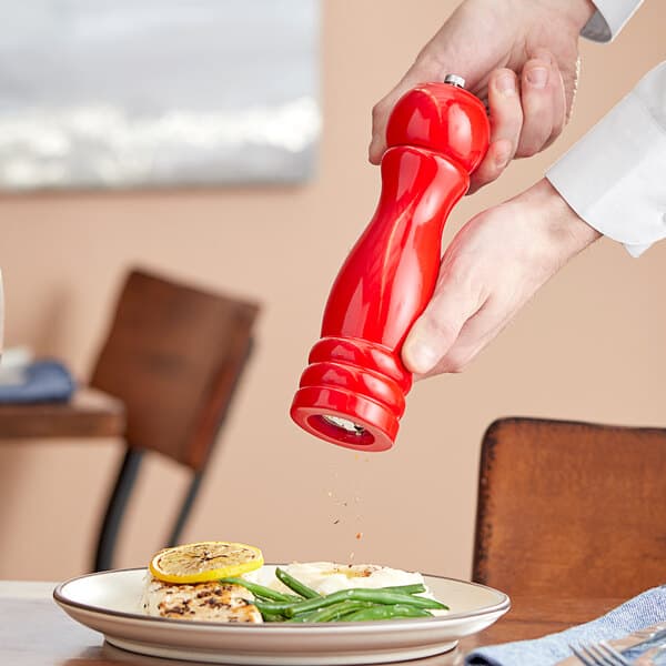 A person's hand using a red Acopa wooden pepper mill to sprinkle pepper on a plate of food.