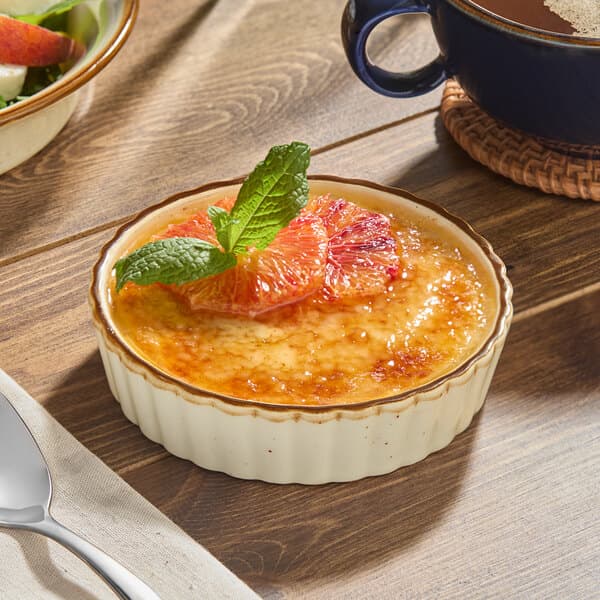 A vanilla bean crème brûlée served in a white stoneware dish, garnished with orange slices and a mint sprig.