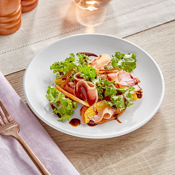 An Acopa matte white stoneware coupe plate with a salad of oranges and greens on a table.