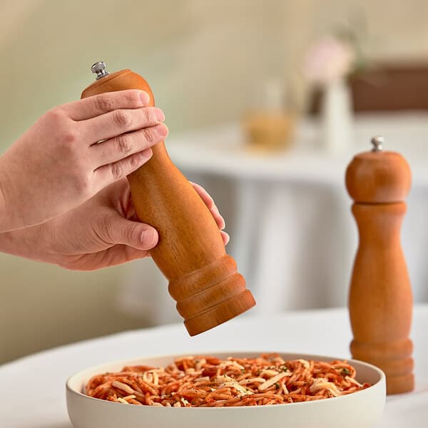 A person using an Acopa wooden pepper mill to grind pepper over a bowl of spaghetti.