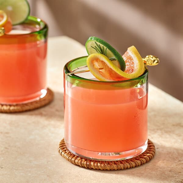 Two Acopa rocks glasses with orange drinks and lime slices on a coaster.