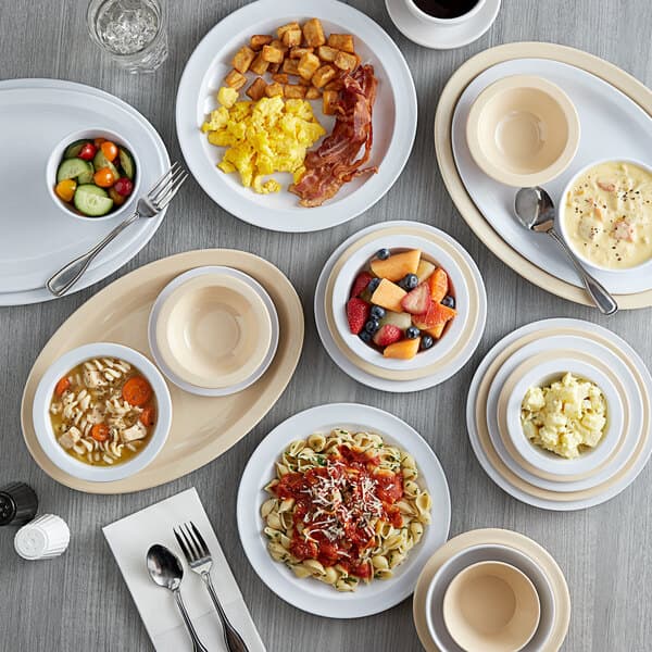 A white table with Acopa white melamine oval platters holding bowls of soup, vegetables, and fruit.