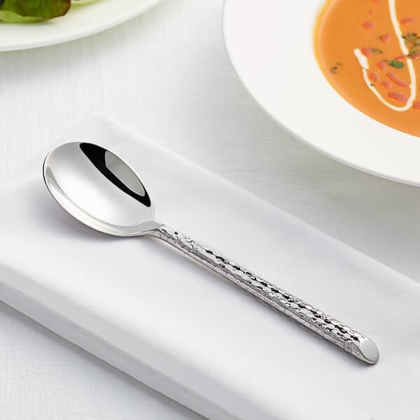 An Acopa Iris stainless steel bouillon spoon on a white napkin next to a bowl of soup.