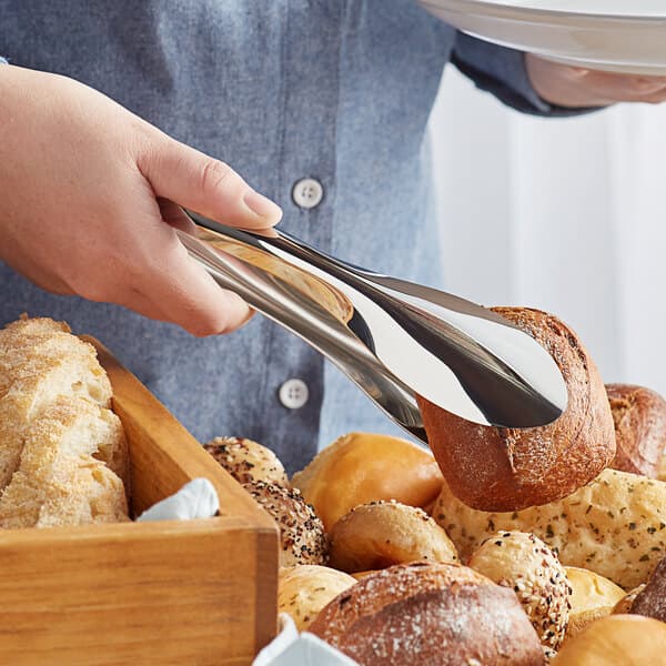 A person using Acopa stainless steel tongs to pick up a piece of bread.