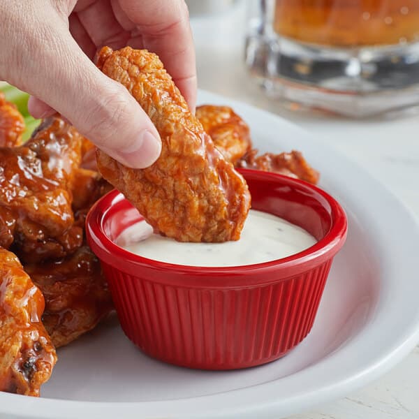 A person holding a piece of chicken dipping it into a red Acopa fluted ramekin of sauce.