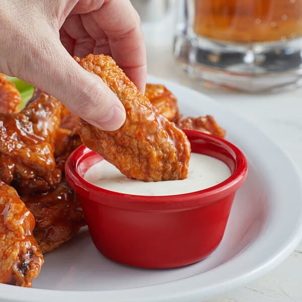 A hand holding a piece of chicken being dipped into a red Acopa ramekin of sauce.