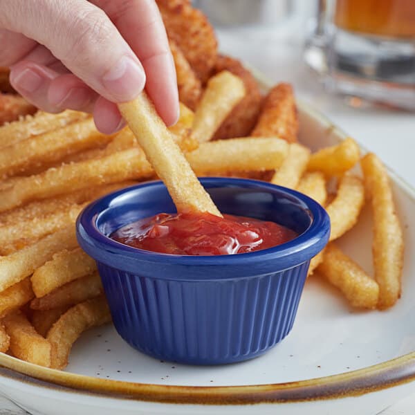 A hand holding a french fry dips it into a blue Acopa fluted ramekin of ketchup.