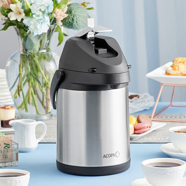An Acopa stainless steel and black airpot on a table with a cup of coffee.