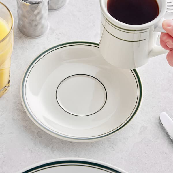 A person holding an Acopa ivory saucer with green bands on a table with a cup of coffee.