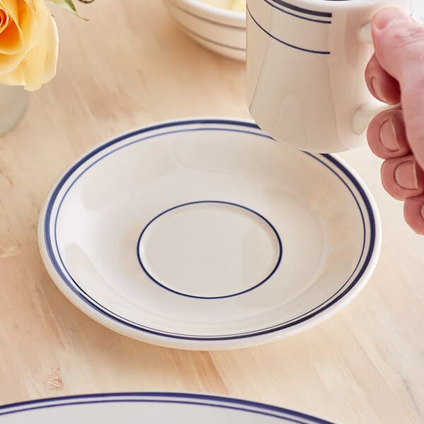 A hand holding an Acopa ivory stoneware cup over a saucer with blue trim.