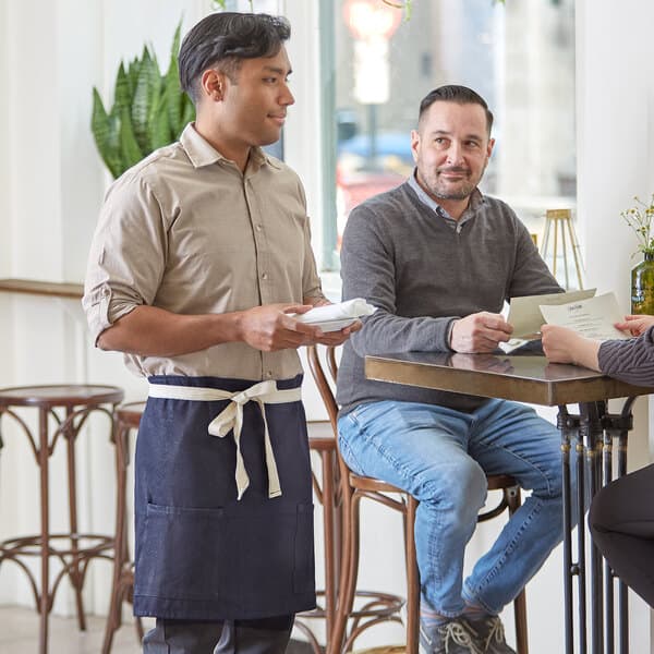 A man wearing a tan shirt and a blue Acopa Kennett half bistro apron serving a woman at a table.