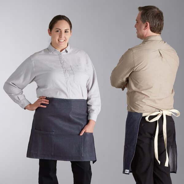 A man and woman standing next to each other, both wearing blue denim half bistro aprons with natural webbing.
