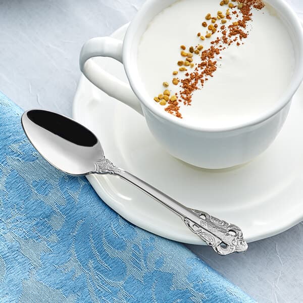 An Acopa stainless steel demitasse spoon on a plate with a cup of coffee and brown crumbs.