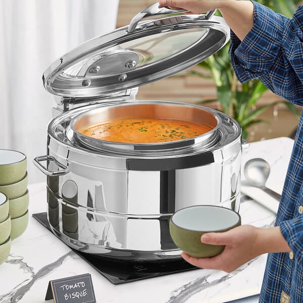 A person holding a bowl of soup in front of a stainless steel chafer.