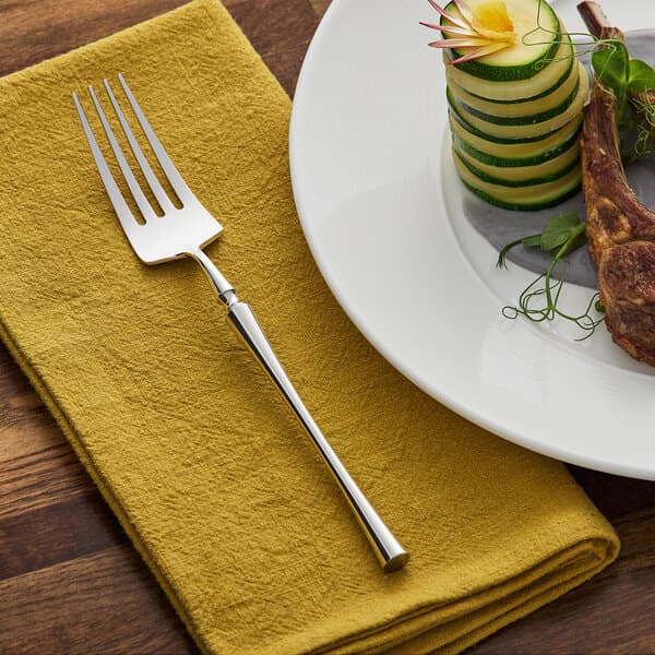 An Acopa stainless steel dinner fork on a napkin next to a plate of food.