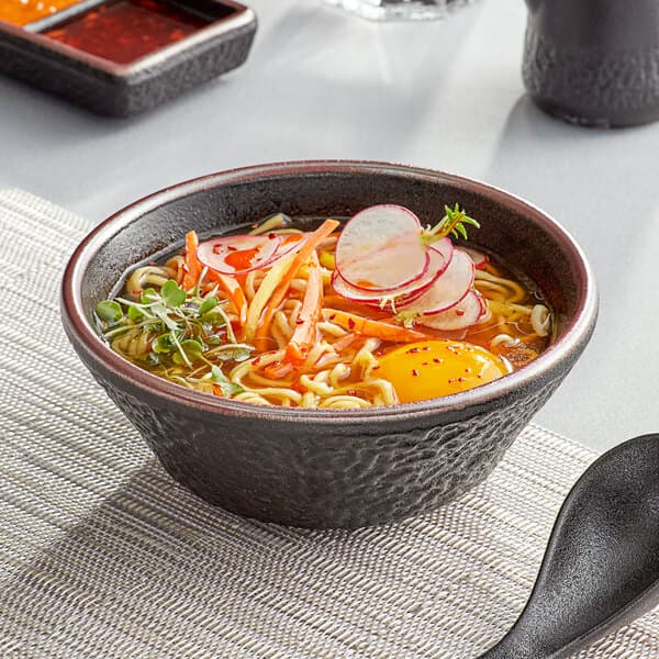 A black Acopa Heika stoneware bowl filled with ramen, vegetables, and an egg with a black spoon.