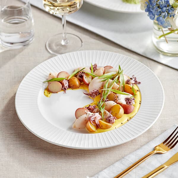 A white Acopa Cordelia porcelain plate with food on a table next to a glass of white wine.